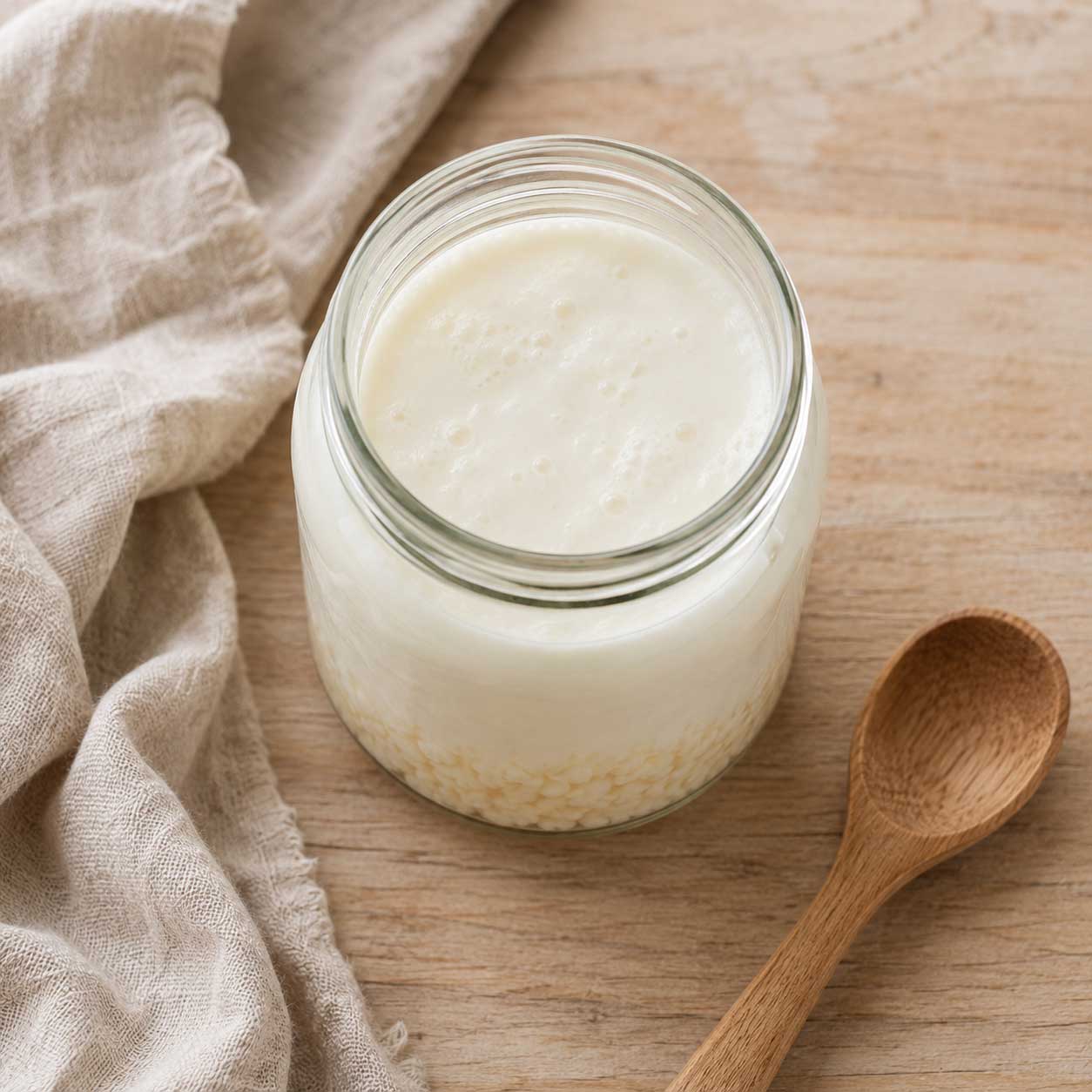 Vista superior de un tarro de cristal con búlgaros de leche fermentando, cuchara de madera y paño de lino al lado